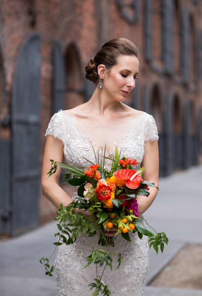 Bridal portrait in cream gown, tropical bouquet with anthuriums and other orange, red and magenta flowers, as well as long greenery that drapes down past brides' grasp