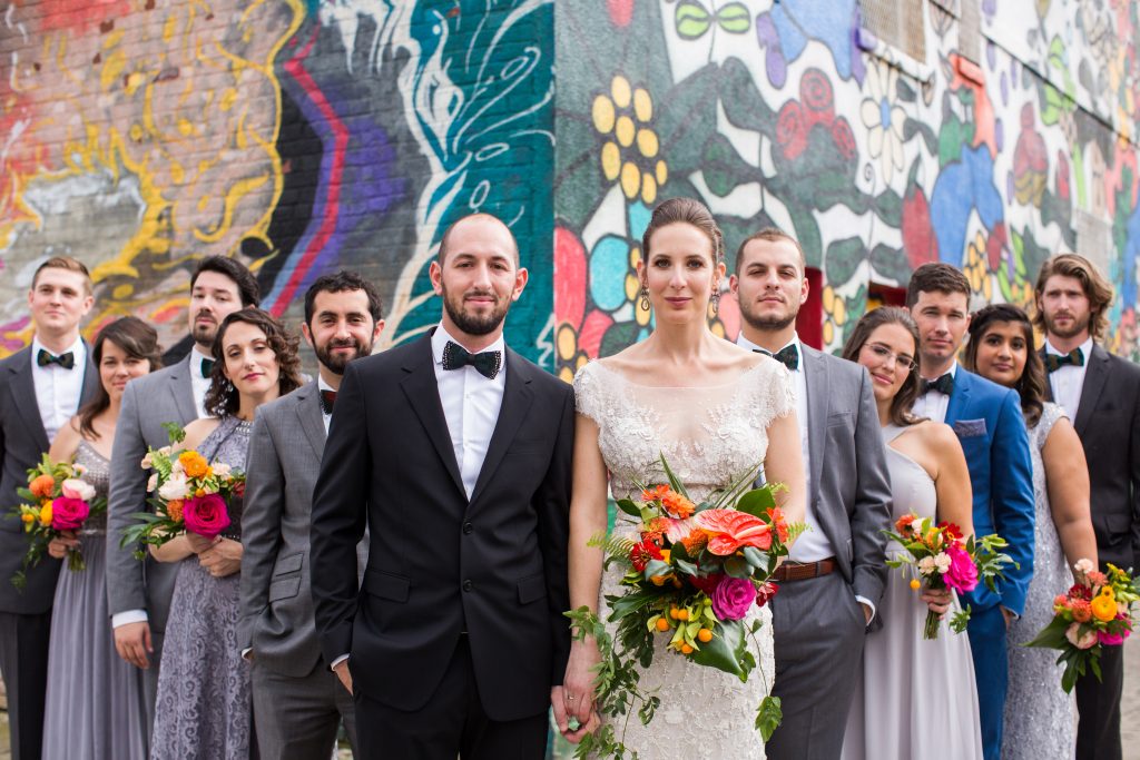 full wedding party portrait, bride and groom front and center. the rest of wedding party fans out behind them in suits of grey, blue or black, grey dresses.