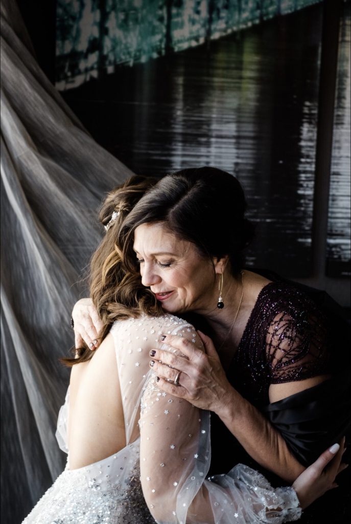 Bride faces away, open back wedding gown and silver sequins. Mother of the bride in a dark beaded dress. She holds her daughter with a bittersweet embrace. Dark background with water.