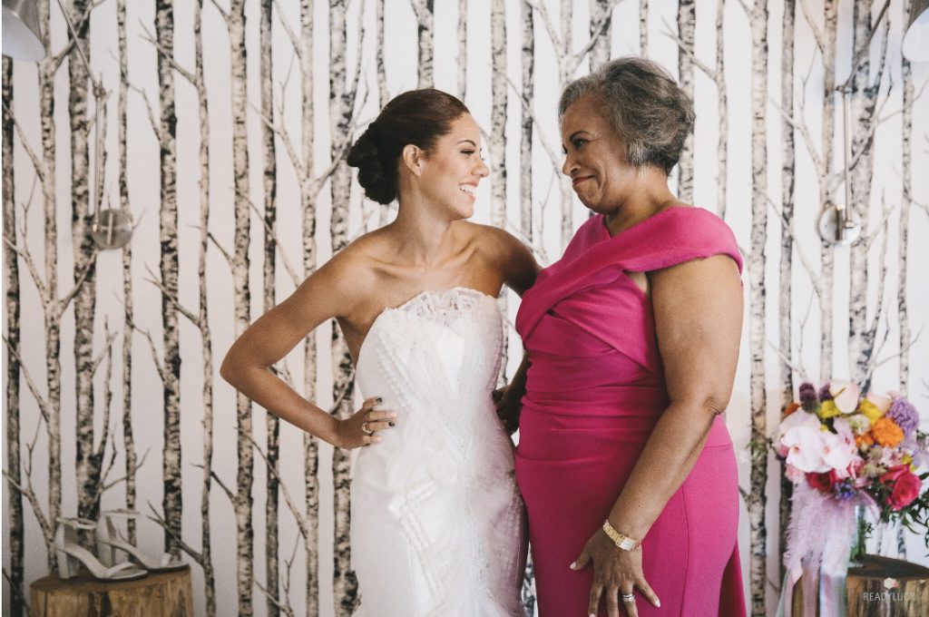 Bride grins at mom on left with updo and strapless, geometric textured white gown. Mother of the bride on the right, fuchsia wrap front dress, smiling at her daughter. 