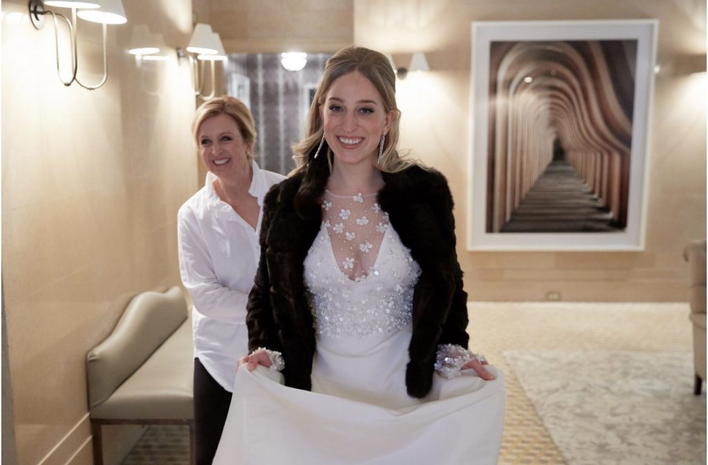 Blonde bride in white gown and black jacket and mother of the bride behind her smiling, hotel room.