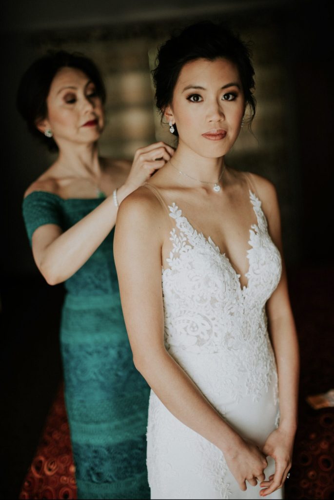 Bride with smokey eyes and white applique dress with transparent straps. Mother of the bride behind in soft focus, with dramatic makeup and red lips, dressed in jade green.
