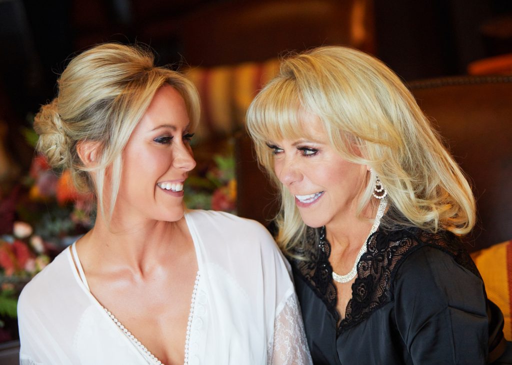 blonde bride in white prep gown, her mother next to her, both looking at each other smiling