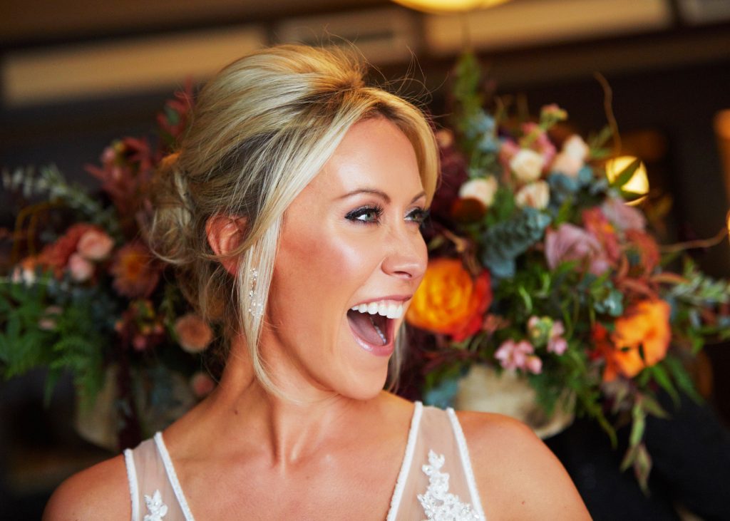Bride looking to the right, blonde hair in glamorous updo, mouth open in big smile. Big bouquet behind her, orange and white blooms with green fronds and ferns.