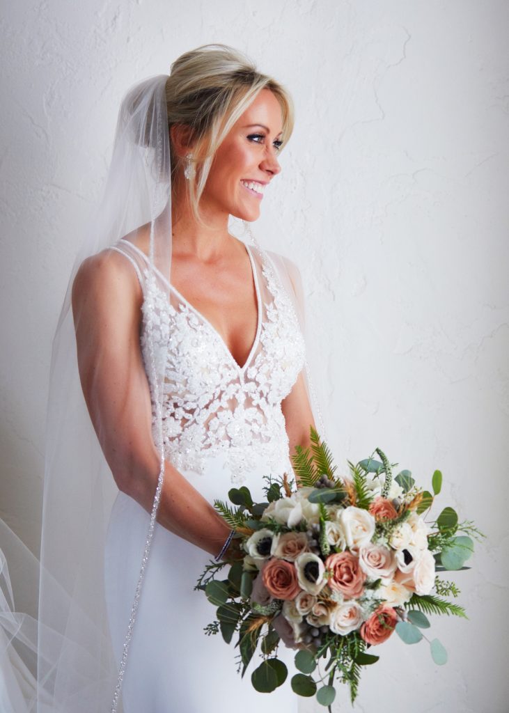 three-quarters portrait of bride in white lacy gown and veil. White and blush blooms of her bridal bouquet with little green drops popping out.
