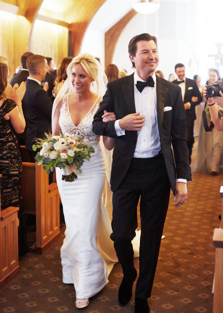 Bride and groom walking down the aisle, white lacy bridal gown and classic black tuxedo. 