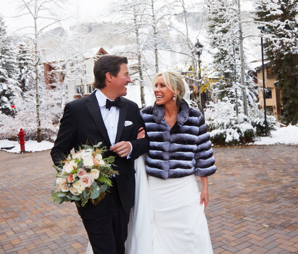 Bride and groom outside in the snow, arm in arm, groom with wedding bouquet in hand. Bride has puffy short jacket on, they're smiling and looking at each other.