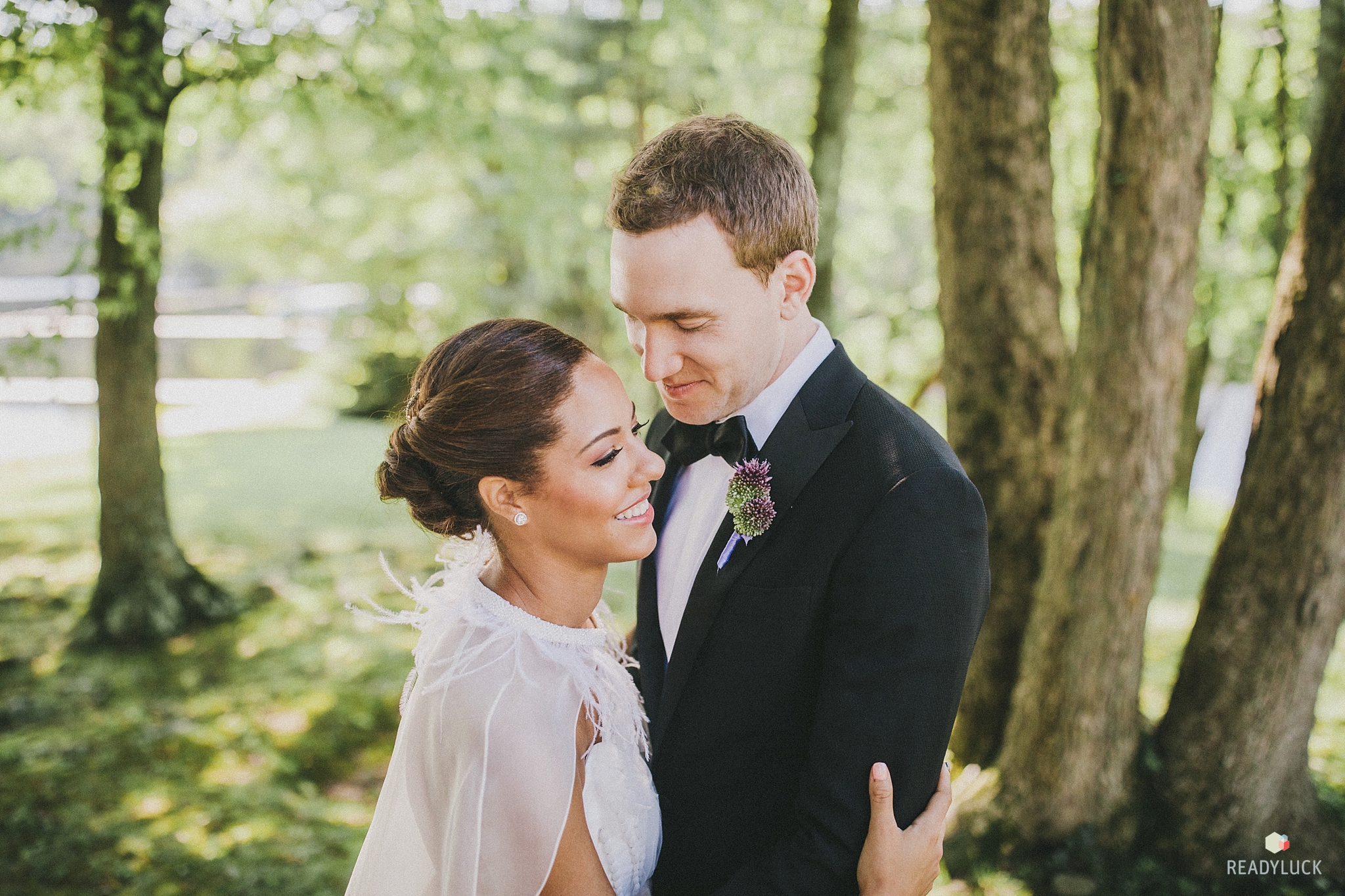 Bride with feathered cape and modern chignon hugging groom on wedding day at Cedar Lakes Estate