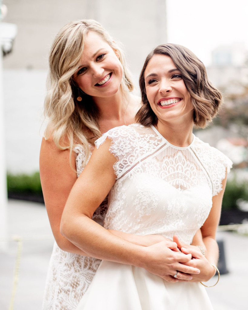 Two brides, one with a dark bob length hair and one with long blonde hair, with modern, edgy textured waves, posing in DUMBO, Brooklyn, on wedding day